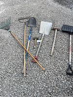 Photo of 7 outdoor tools laid out on gravel, including rakes, shovels, a sledgehammer, and a broom.
