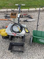 Photo showing push spreader with pressure washer gun and hose mounted, along with long-handled gardening tools and black rectangular container on gravel surface.