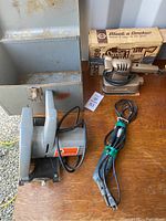 Three Black & Decker power tools: finishing sander with box, rotary tool, utility saw on wooden table