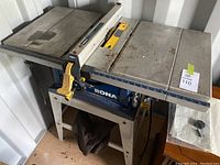 Front and side view of the Rona table saw and stand, showing the overall structure and condition including the fence guide and blade guard.