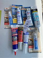 Lot of 8 silicone sealant tubes arranged on a surface. Various brands including Permatex, DAP, LePage, GE, unopened condition.