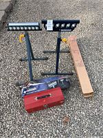 Two metal support stands with roller tops, red metal toolbox, and boxed ratchet straps on gravel surface.