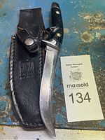 Curved fixed blade knife with black handle and leather sheath on blue surface with auction card