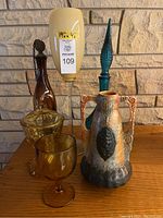 Full lot view showing clay vase, brown glass decanter, blue glass decanter, amber goblet, and amber covered dish on wood surface against brick background