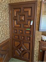 Tall wooden door with dark brown finish and detailed raised geometric panel design, one side with a metal round door handle and brass hinges, set against a floral wallpapered wall.