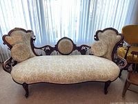 Full front view of carved wood frame settee upholstered in cream and gold toile fabric, with two round pillows on seat.