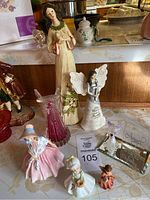 Seven assorted figurines displayed on a table including angel figures and Royal Doulton Lily. Figurines include a tall angel in cream with green collar, a white angel holding a silver object, Royal Doulton Lily in pink dress and bonnet, plus smaller angels and decorative figures.