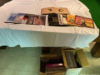 Two large boxes filled with assorted vinyl records shown on and under a table with white cloth, displaying multiple record covers and record sleeves.
