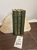 Charles Dickens Complete Works three-volume set with green leather spines and gold embossing placed between two stone bookends on a textured surface, includes lot tag.