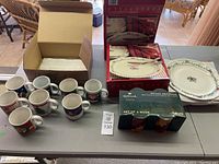 Photo showing 12 Christmas mugs and 8 plates on table, some packaging visible