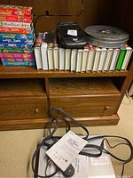 Photo showing 27 Disney children’s VHS movie cases stacked in two rows mainly Winnie the Pooh titles, a Mickey Mouse tin, a VHS rewinder device, and a 16mm film reel canister on a wooden shelf.