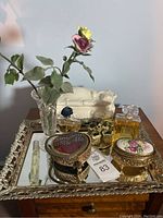 Overview of mirrored tray holding vintage dressing table items including two ceramic floral box tops, two perfume bottles, heart-shaped brass trinket box, small vase with artificial flowers.