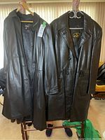 Two men's black leather trench coats hanging side by side, one with button closure and smooth leather, the other with button closure, belt and slightly textured leather.