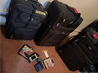Three black soft-sided suitcases standing upright with travel accessory items laid on the floor in front, showing the overall lot contents including tags and packaging.