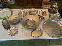 Photo showing twelve silverplated and metal decorative pieces arranged on a table including trays, hand-forged bowls, tiered tray, and small items