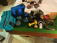 Photo showing five 5 lb bowling balls, two pairs of gloves including a Rawlings glove, one or two sports bags, a pair of yellow small hand weights, and some smaller items on a green felt surface.