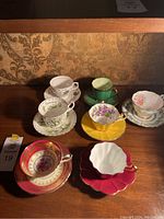 Seven assorted vintage teacups and saucers arranged on a wooden surface with vintage wallpaper background, showing variety in color and floral designs.