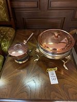Two vintage copper pots on a wood table. One pot on a stand with three curved legs and a burner underneath, the other smaller copper pot with a vented lid and a handle.