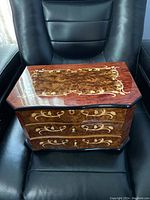 Jewelry box closed front view sitting on black chair showing burl wood inlay and brass floral designs with three drawers.