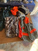 Photo showing hunting vest and pants in camouflage, orange safety vest and caps.