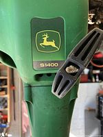 Close-up photo of the John Deere logo and S1400 model number on the green trimmer housing, showing some dirt and wear.