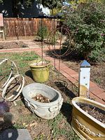 Photo showing three ceramic planters: green round, white rectangular with lion heads, and yellow-tan oval planter, along with metal tomato cages and decorative orbs in background, plus garden stakes visible.