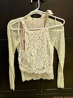 Two white lace tops hanging side by side on white hangers against a dark background. Sleeveless lace top with shoulder ties in front, long sleeve lace blouse behind it.