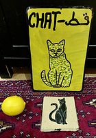 Front view of yellow metal sign with a hand-painted cat image and Arabic text, shown along with cat keychain on textile surface