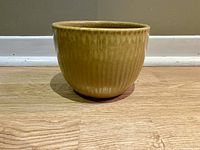 Single medium-sized pottery pot with creamy yellowish glaze and vertical ridges against wooden floor and wall baseboard