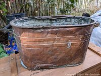 Large old copper boiler with dents and patina, photographed outdoors on a wooden surface from a side angle showing its oval shape and side handle.