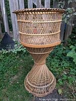 Full view of vintage wicker and bamboo plant stand outdoors against a wood fence, showcasing its hourglass shape and woven details.