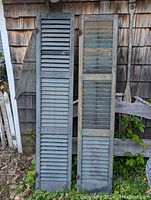 Pair of antique green wood shutters leaning against a shingled wall, showing full length and original hardware.