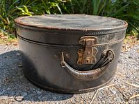 Front wide-angle view of the black round antique suitcase showing leather handle and metal lock hardware with no key.