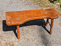 View of early pine bench showing curved top and four legs on paved surface outdoors.
