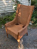Side angle view of the early wicker chair placed outdoors on gravel in front of siding, showing the weave detail and straight legs.