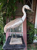 Side view of carved wooden heron sculpture on black surface with rustic background, showing overall shape, steel legs and wood grain.