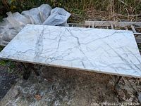 Full view of rectangular white marble top coffee table with grey veins resting on wrought iron legs outdoors.