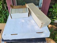 Photo showing two long cardboard boxes containing large quantities of trading cards stacked neatly inside. Cards are standard size and cover hockey and baseball themes.