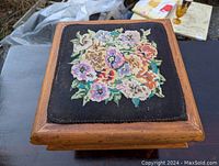 Top view of the needlepoint footstool showing the detailed floral basket design on black fabric with wooden frame.