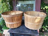 Two small round wooden apple baskets, 14 inches across, made with split weave ash wood, placed outdoors on a black table with plants in the background.