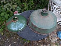 Two large green enamel-coated metal barn shades placed on a black table outside, one bigger and one smaller, both showing surface rust and wear.