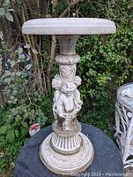 Front view of cream-colored cherub plant stand with a round top and detailed column with cherubs.