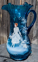 Photo of large blue glass water pitcher with flared rim and handle, featuring a hand painted Mary Gregory style woman in a white dress and white ferns on the body.