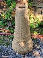 Full view of the tall tapered unglazed beige ceramic plant stand standing outdoors on gravel and wood with some grass around.