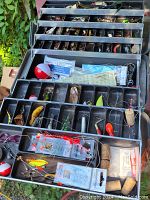 Open tackle box showing multiple trays filled with a wide assortment of fishing lures, bobbers, hooks, and packaged fishing accessories