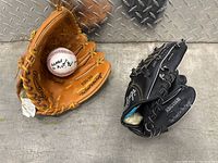 Two Rawlings baseball mitts, one tan and one black, with a signed baseball inside the tan mitt on a concrete surface.