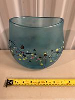 Blue glass vase with decorative abstract dots and curved top, shown with measuring tape indicating about 7 inches wide.