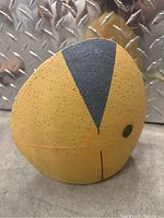 Front view of a small yellow ceramic art piece shaped like an abstract bird with painted black top and green eye detail laid on a concrete surface with a metal diamond plate background.