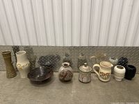 Wide image showing all 10 ceramic items arranged in a row on a metallic surface with diamond plate background. Items vary in shapes, glazes, and decorative motifs.