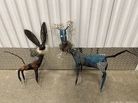 Two handcrafted animal sculptures placed side by side on a concrete floor against a white and metal corrugated background.